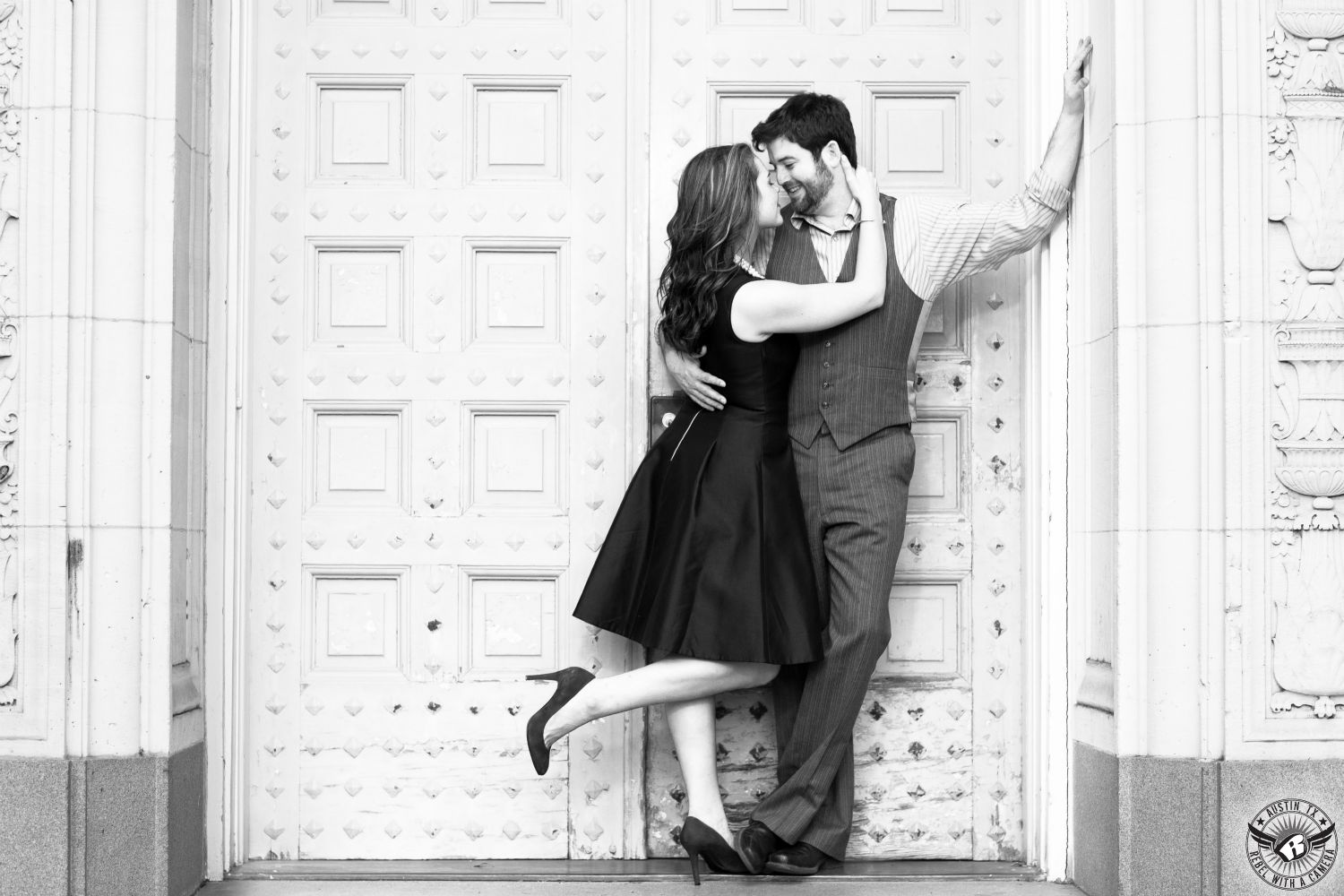 brunette girl in black dress with black heals hold a guy wearing a grey and white vest and pants tightly in front of a studded vintage  door at UT in this engagement pic in austin